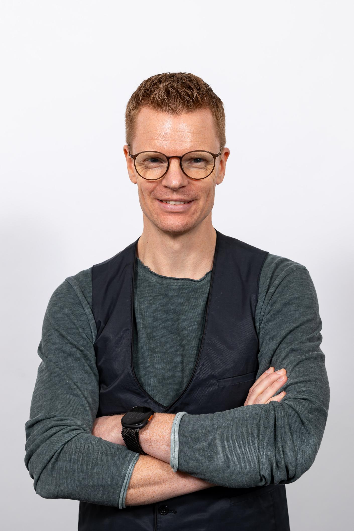 Portrait of Stefan Mecke. Portrait of Stefan Mecke. He is wearing a gray sweater and a black vest and is smiling at the camera.