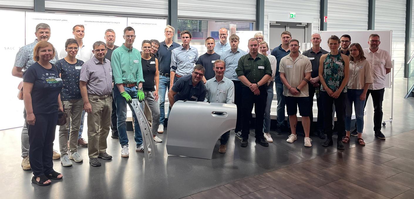 The interdisciplinary Car2Car project team, comprising experts from industry and academia, is pictured in a modern industrial hall; a silver-colored car door is in the foreground.