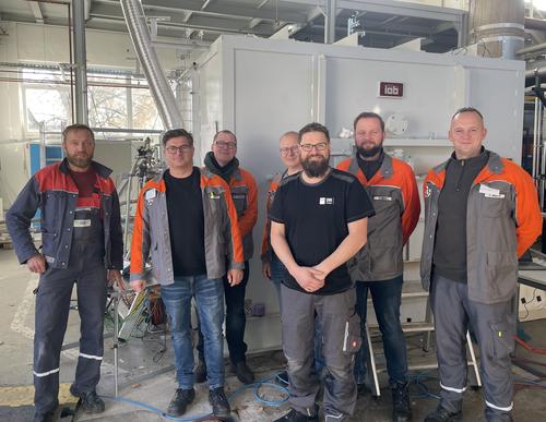 Das Projektteam. Sechs Mitarbeitende stehen für ein Gruppenfoto vor einer großen Industrieanlage in einer Werkhalle. Vier tragen orangefarbene Arbeitsjacken oder Warnkleidung; zwei stehen in dunkler Kleidung in der Mitte.